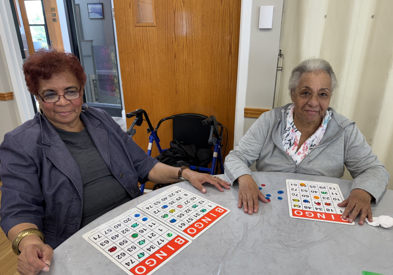 Two older women sit at a table with bingo boards. They smile at the camera.
