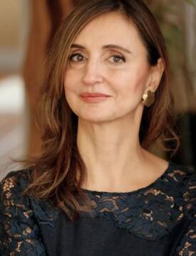 A middle-aged woman with long brown hair, brown eyes, looks to the camera. She wears a black top with lace