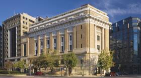 View of the National Museum of Women in the Arts building from outside showing the Neoclassical building from one corner. The building is a tan-colored stone with an arched doorway, long vertical windows, and detailed molding around the roof.