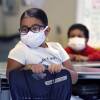 A girl seated at a classroom desk twists around to look behind her. She and another child, out of focus in the rear, are both wearing masks.