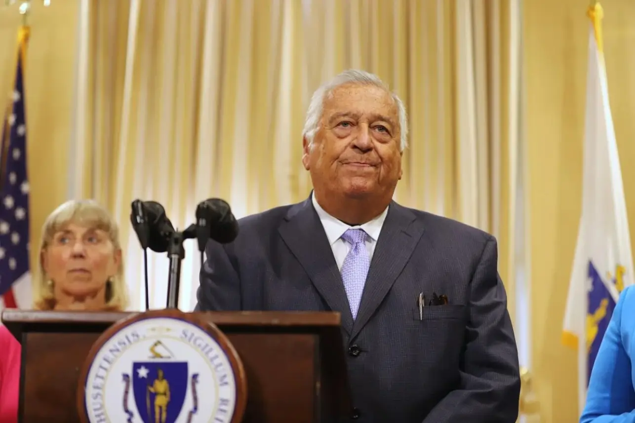 A man dressed in a suit and tie stands behind a podium with a microphone in the foreground as a woman glances off-camera in the background.