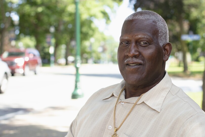 A black man with short cropped grey hair and mustache poses for a portrait sitting on a street-side bench on a sunny day.
