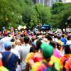 Hundreds of people wearing colorful clothing and waving rainbow flags walk down the middle of a city street, with fluffy green trees on one side and apartment buildings on the other.