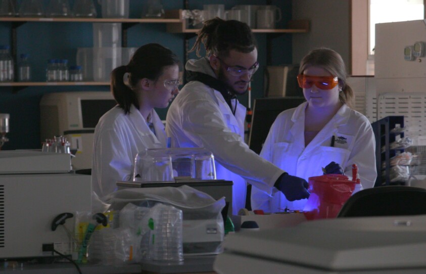 3 young people in white lab coats gathered in a lab with little lighting looking at equipment that emits a purple glow