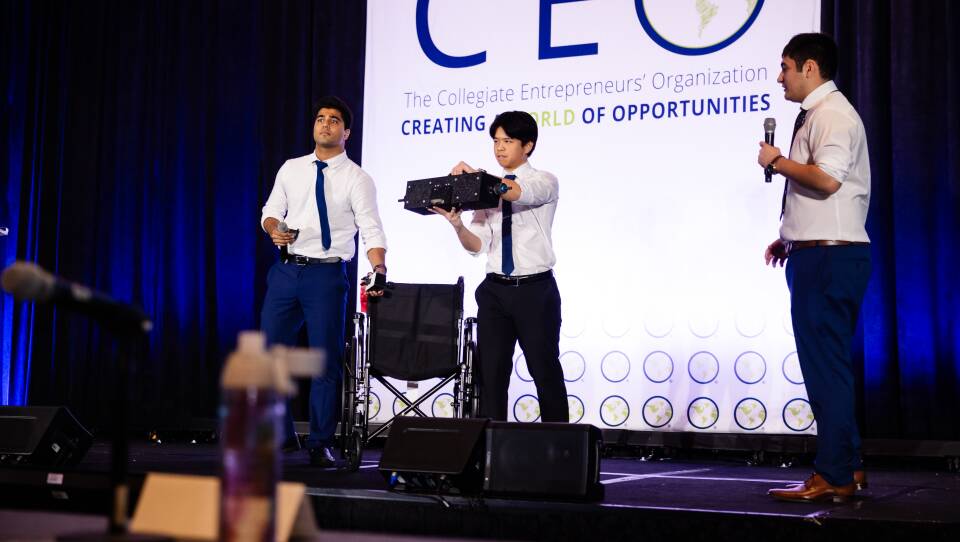 Three young men in buttondowns, ties and slacks presenting a pitch with a wheelchair onstage.