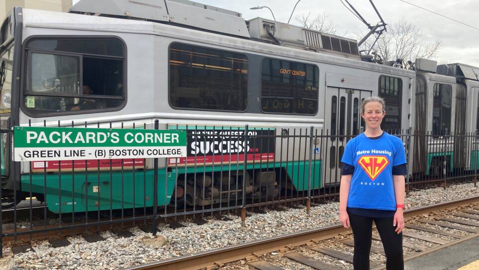 A woman in a shirt reading Metro Housing Boston stands in front of train tracks. Behind her, a Green Line trolley passes on the tracks. A Packard's Corner station sign is visible.