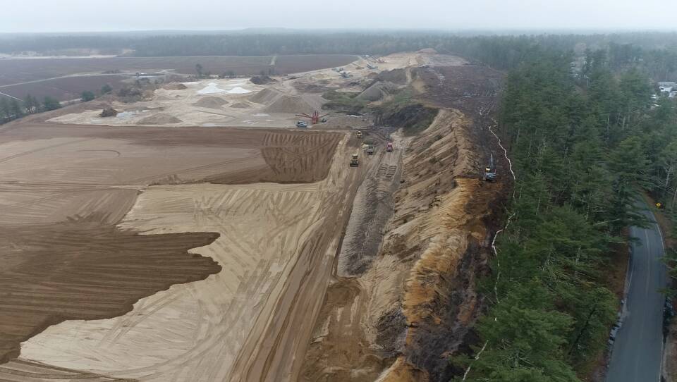 A large cleared area with sand and dirt that has a few pieces of machinery. On the right is a road with a thin strip of evergreen trees separating the road from the excavated area.