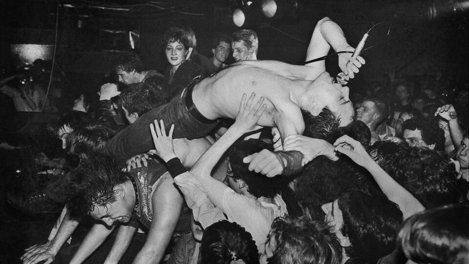A black and white image of a shirtless man crowdsurfing while singing into a microphone.