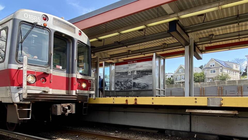 A subway train with a broad red stripe across the front pulls into an above-ground station on a sunny day.