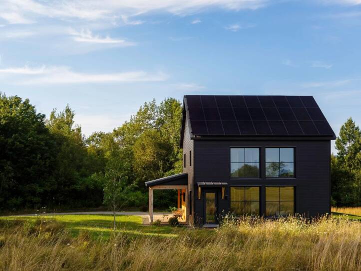 A passive house sits in a green field surrounded by trees and tall grass. The exterior is black and it has four large windows on the side.