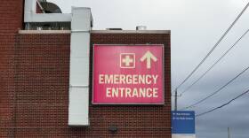 A red sign is posted on the side of a brick building. It says "emergency entrance" and has an arrow pointing forward.