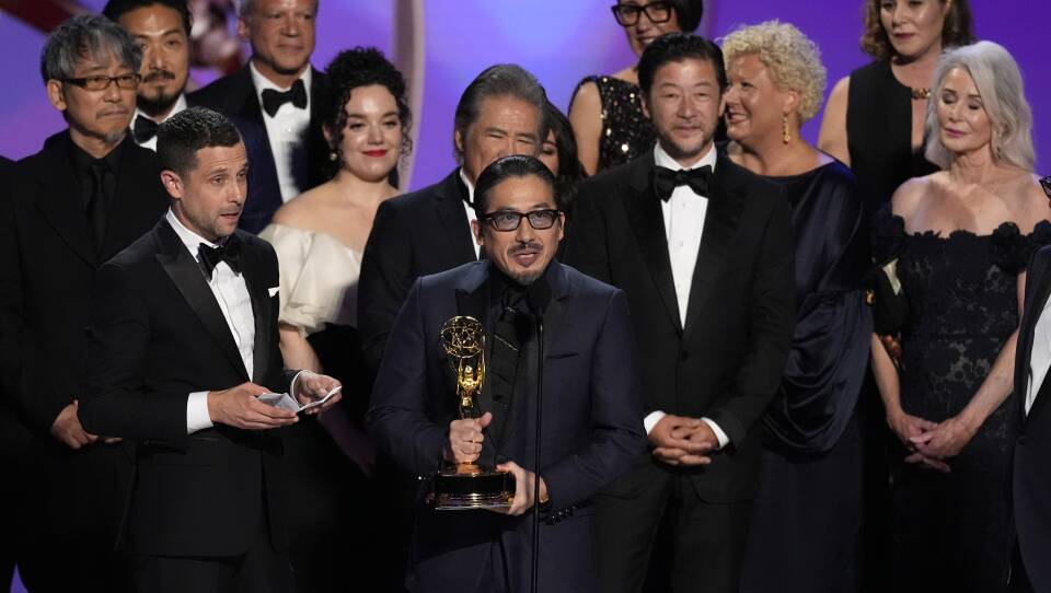 A group of people in suits and gowns stands on a stage behind a man holding a golden award statuette.