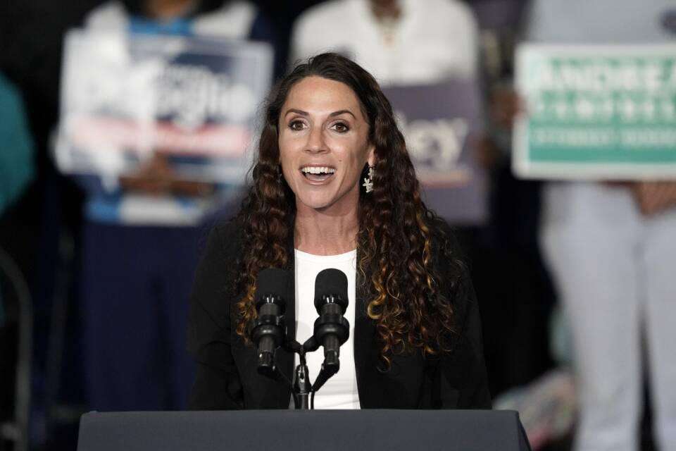A woman with long curly hair speaks as she stands behind a podium with microphones on stage, with people holding political campaign signs behind her.