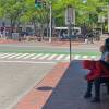Two people sit on a bench in the shade of a street corner by a bus stop sign. Another person stands near them.