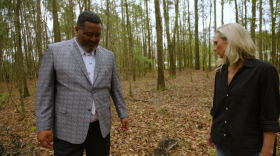 A Black man and white woman stand on a piece of land, covered in trees. They're both looking down at the ground.
