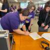Three women lean over a document on a table as the woman on the left signs it. Two of them wear purple union shirts.
