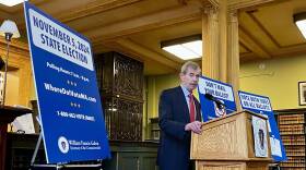 A man in a blue suit stands behind a wooden lectern, flanked by blue signs bearing advisories about the Nov. 5, 2024 election.