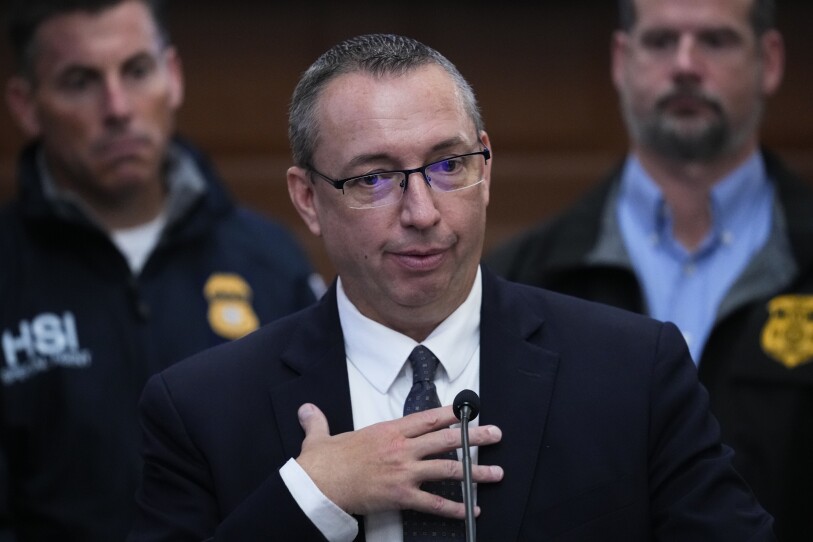 A man in a suit and tie holds his hand over his chest as he speaks into a microphone at a press conference, with police in uniform behind him.