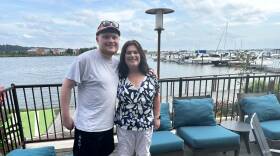 A young man and woman pose and smile for a photo in front of a marina. He has his arm around her shoulder.