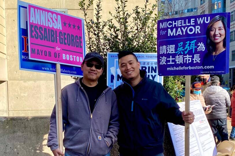 A man in a gray hooded sweatshirt is pictured on the left holding a pink sign for Boston mayoral candidate Annissa Essaibi George, while a man in a blue jacket stands on the right holding a sign for her opponent, Michelle Wu.