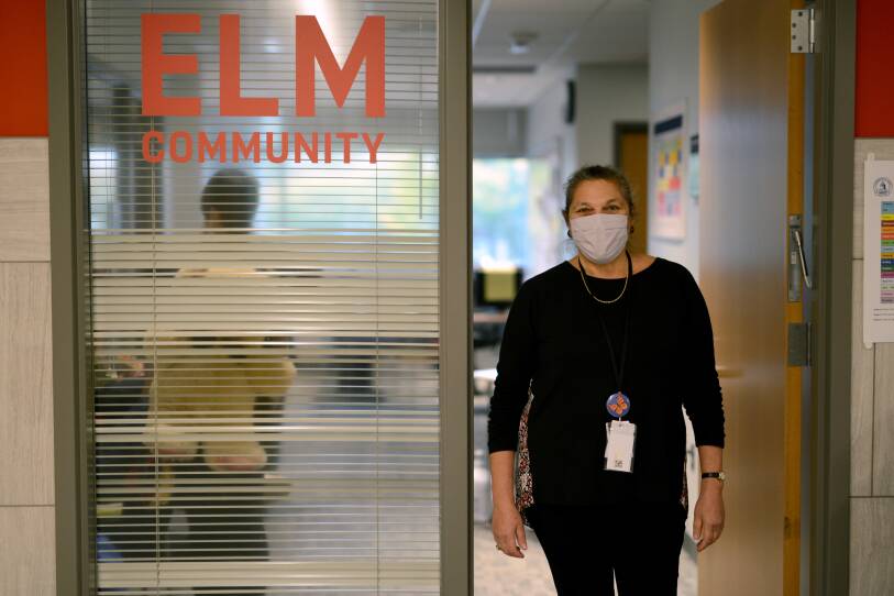Davila, wearing a black shirt and pants, stands in the doorway of an office. On the wall next to her, orange text reads "ELM COMMUNITY."