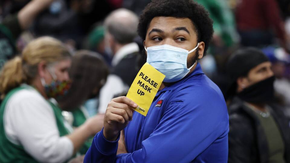 A man wearing a mask holds up a yellow card that reads "Please mask up" as a crowd of people walk around him.