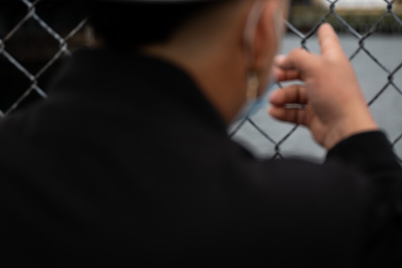 A young man wearing a surgical mask points through a chain-link fence