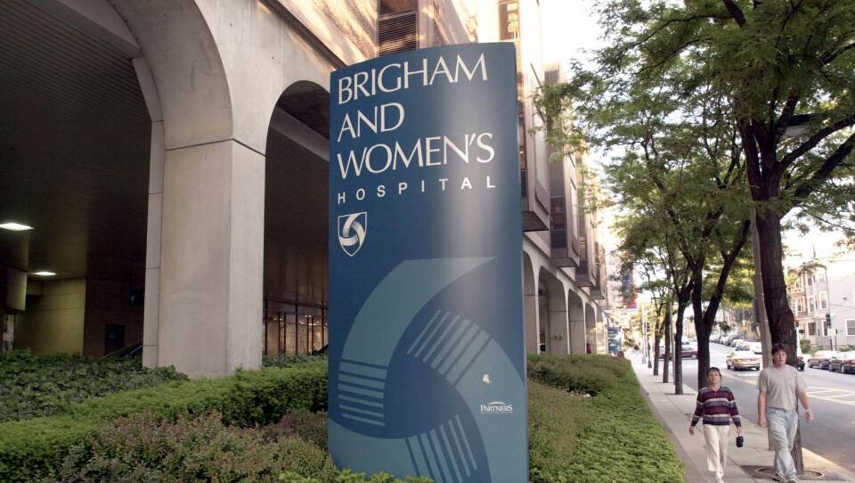 Two people walk on the sidewalk past a large blue sign that says "Brigham and Women's Hospital"