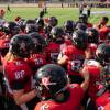A group of women gather on a football field. They wear black football helmets and red jerseys.