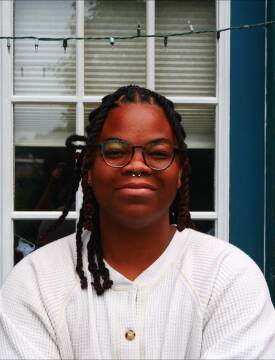 A person wearing glasses and braided hair smiles slightly in front of a door.