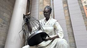A massive bronze statue of a Black woman, seated and holding a birdcage that she's opened. A free bird sits on top of the bird cage, staring at the woman, who wears a simple white dress of stone or plaster.