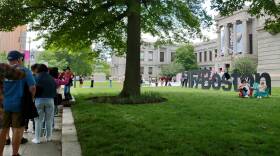 A line of people stand on the sidewalk around a large museum while others sit in chairs and pose by the MFA Boston sign in the lawn.