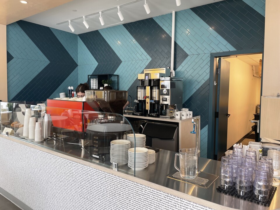 This is the interior of a cafe, in the background are coffee, espresso and cappuccino machines. In the foreground is a stainless steel countertop covered with plates and coffee cups.