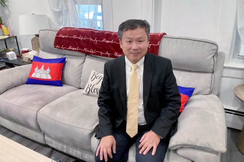 A man in a suit and yellow tie sits on a gray couch. The couch is topped with a red blank and two of its three pillows have the image of the Cambodian flag.