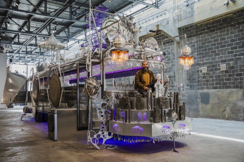 This is a photograph of a large sculpture. The structure is mostly made out of metal. It looks like a combination of a bus and a locomotive. sitting at the front of the structure, as if he were in the driver's seat, is the artist who made it.