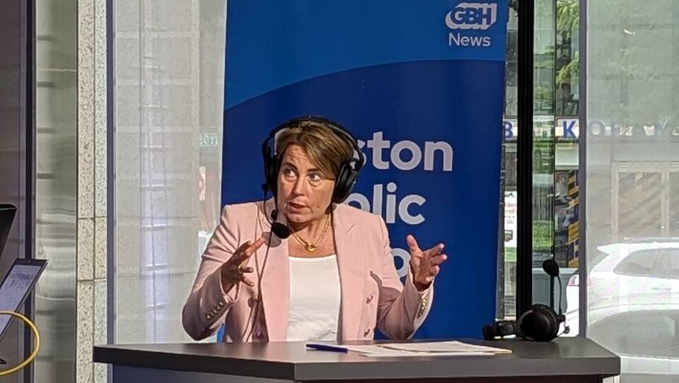 Gov. Maura Healey gestures with her hands on the radio set. She wear a pink blazer and radio headset.