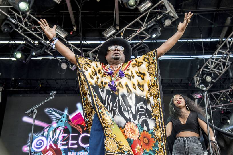 A man wearing a colorful outfit stands on stage with his arms up in the air.