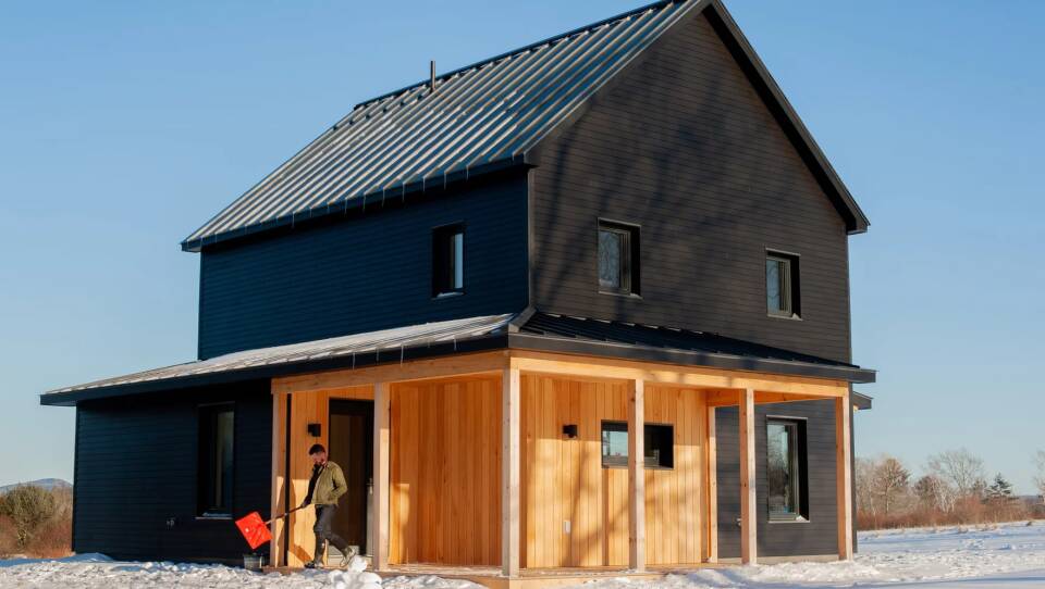 A passive house stands in an open lot, it is wooden and black. A white man with a shovel walks through the snow out front.