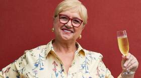 A woman wearing red-framed glasses and a floral shirt smiles as she holds a glass of champagne as if participating in a toast.