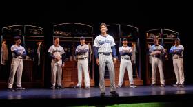 This photo is from the play, "Take Me Out."  On the stage are seven baseball players. They are in the locker room, still in their uniforms. Standing apart from his six teammates is one ball peers who stands at the edge of center stage. He is looking outward. The expression on his face looks like one of frustration.
