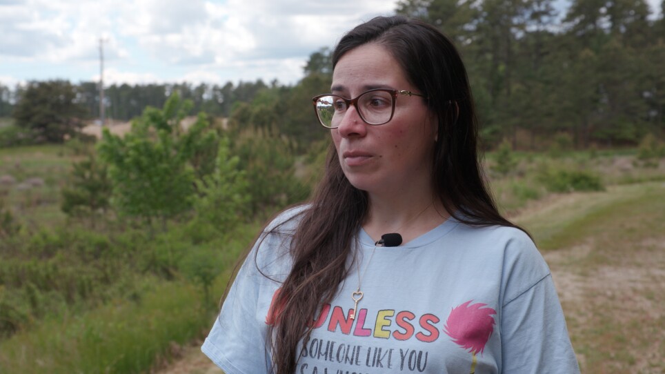 A woman with long brown hair and glasses stands outside.