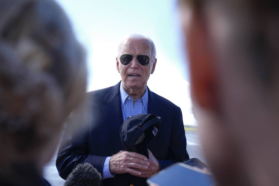 Joe Biden wears sunglasses and holds a light jacket in his hands as he speaks outside at an airport.