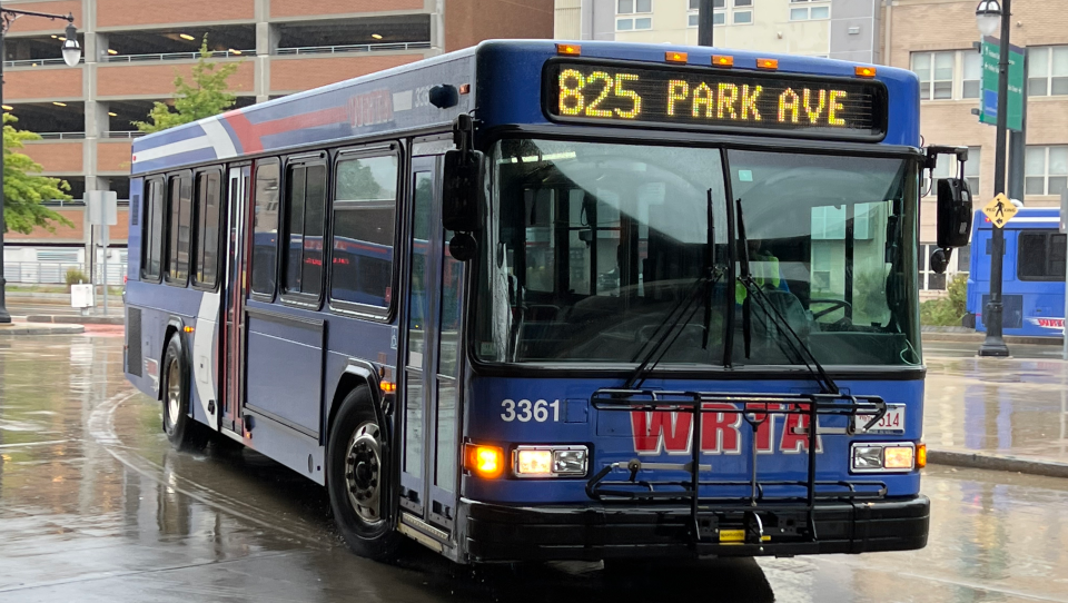 A blue bus turns around the corner of a city street on a rainy day.