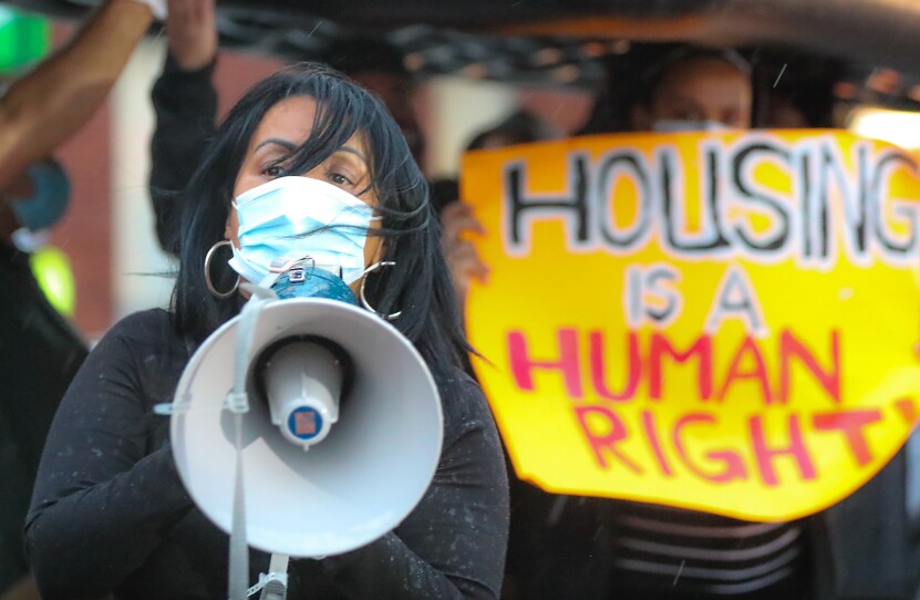 A woman speaks into a megaphone in front on someone holding a yellow sign with the words "housing is a human right" displayed in black and red letters.