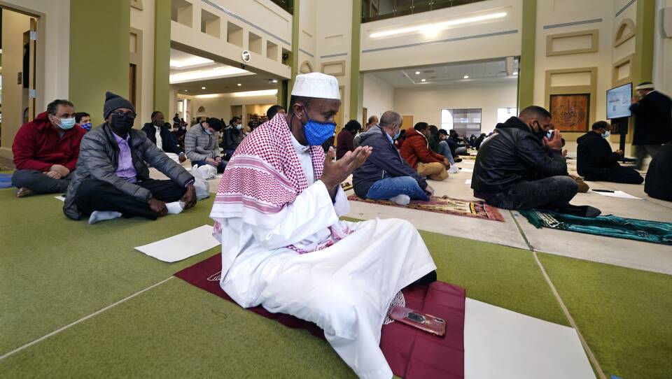 A man sits on a mat with his legs crossed as he holds his hands in prayer. He is in a large room with other worshipers.