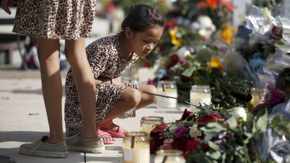 A young child wearing a dress and sandals squats in front of candles and flowers