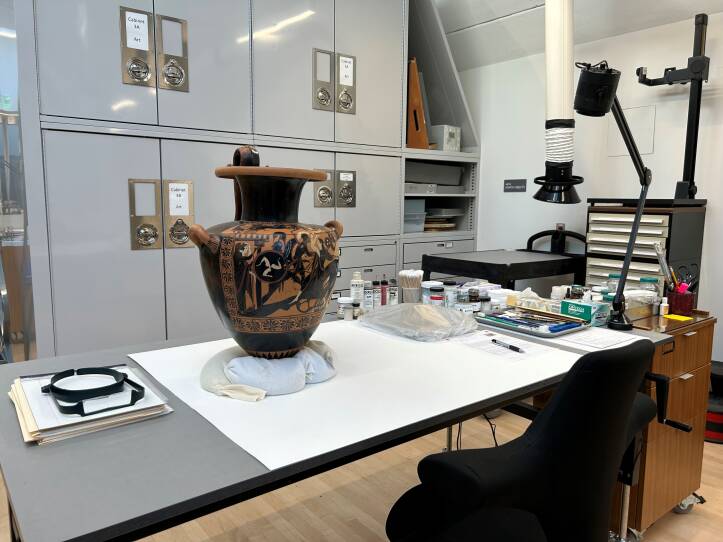 A large brown and tan urn decorated with human figures sits on a table, held still by sandbags, in an art workshop.