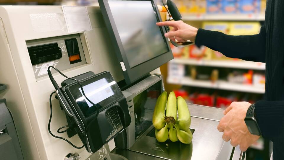 A person selects a button on the screen of a self-checkout kiosk as they weigh bananas on the scale.