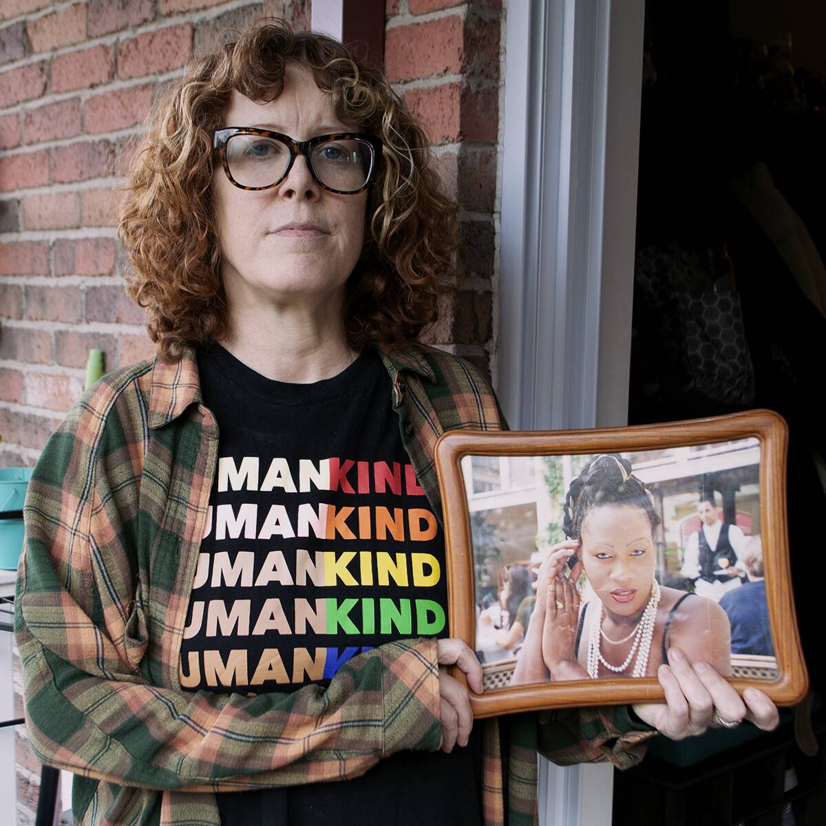 A woman stands outside the front door of an apartment, holding a framed photograph of another woman.