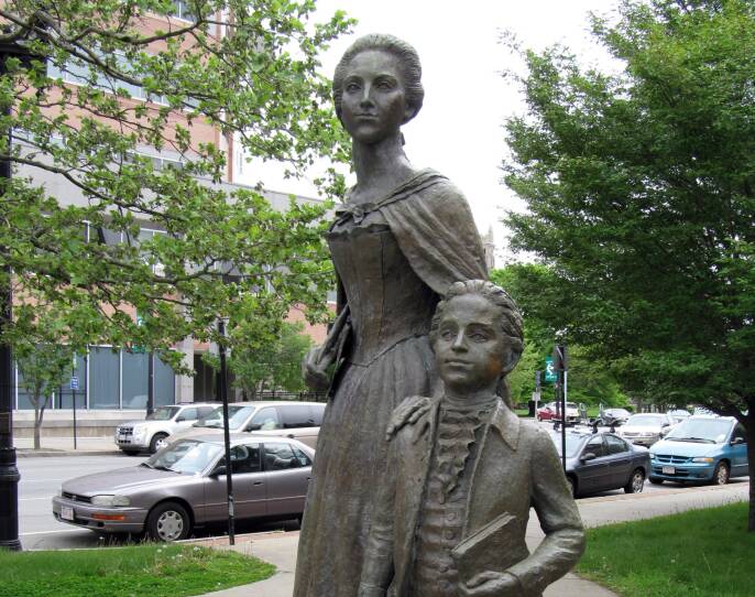 A bronze statue shows two figures: a woman in a dress with a young boy who's holding a book. Both look off pensively in the same direction.